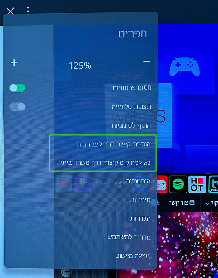 מסך טלוויזיה המציג תפריט הגדרות בעברית עם אפשרויות הקשורות לגודל תצוגה והגדרות כפתור הבית. סמלי אפליקציה שונים נראים חלקית ברקע.